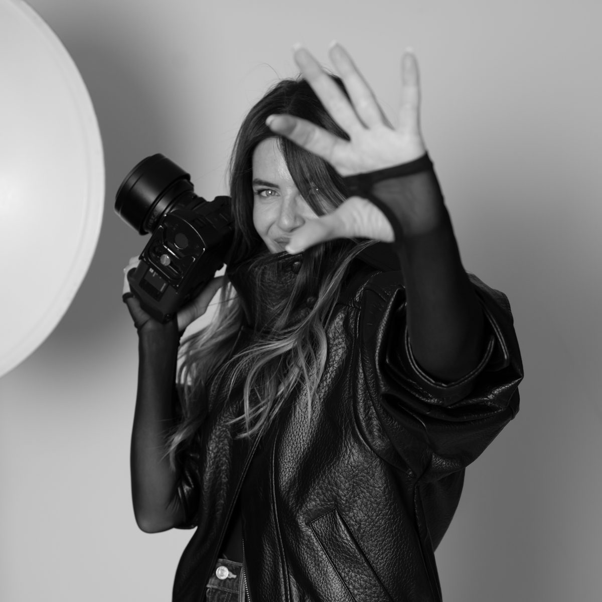 Shiran Itzhaki holds up her camera and raises one hand toward the lens, in a black and white studio portrait.