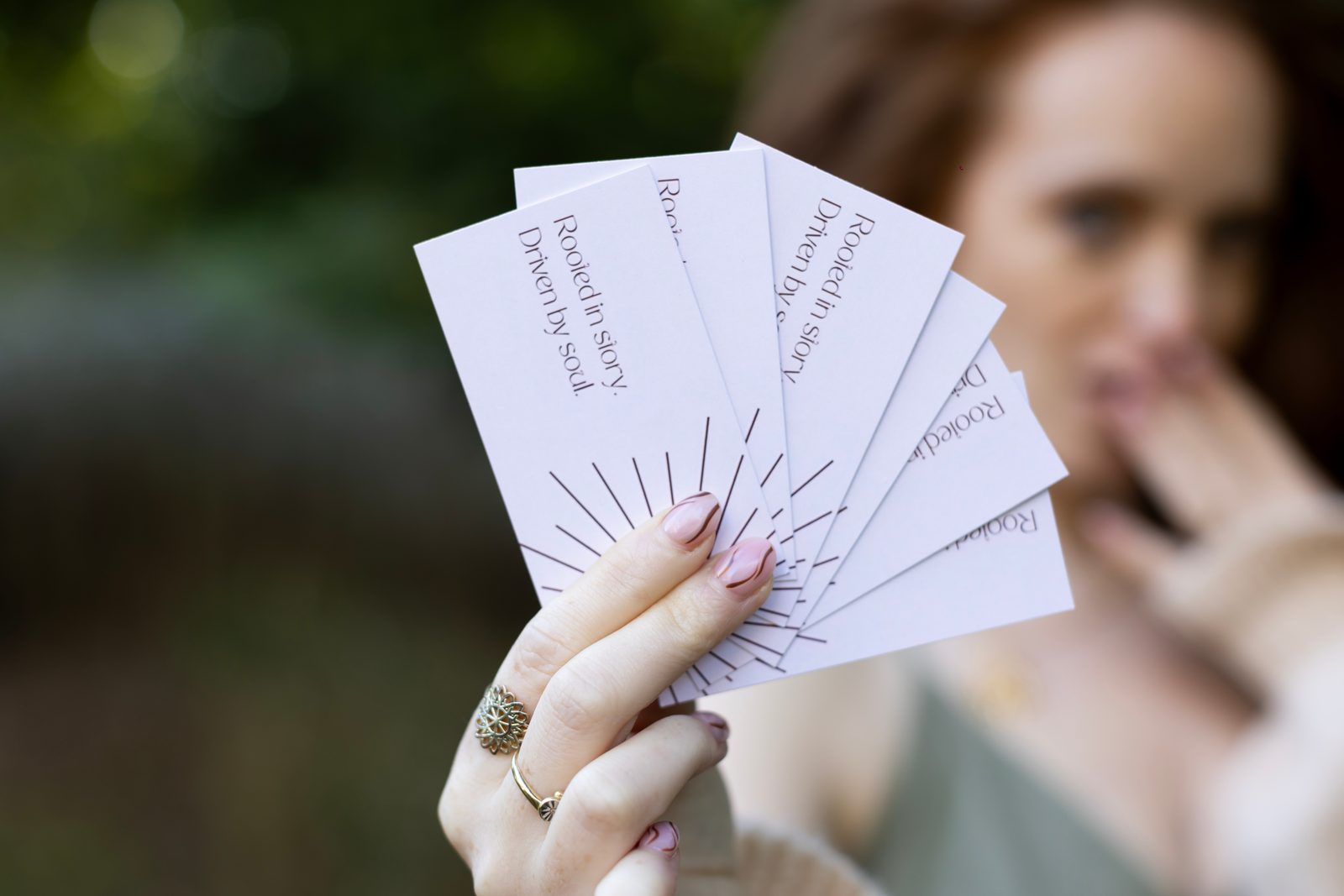 A hand holds a fan of printed business cards, with a woman out of focus behind them.