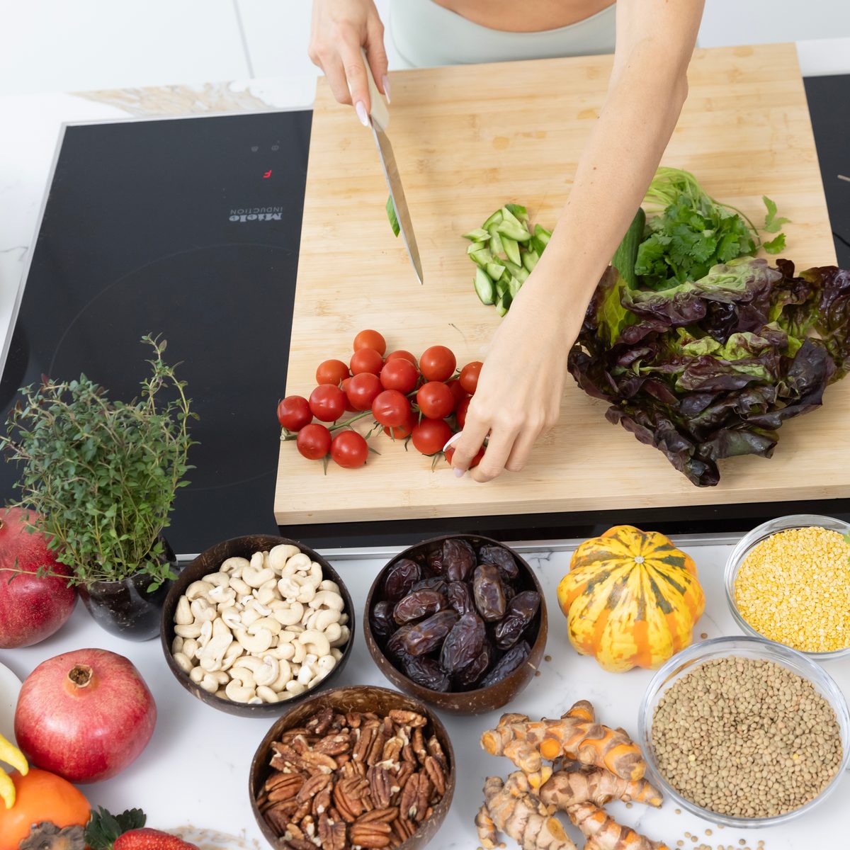 Hands slice vegetables on a wooden board, surrounded by bowls of nuts, grains and fresh produce.