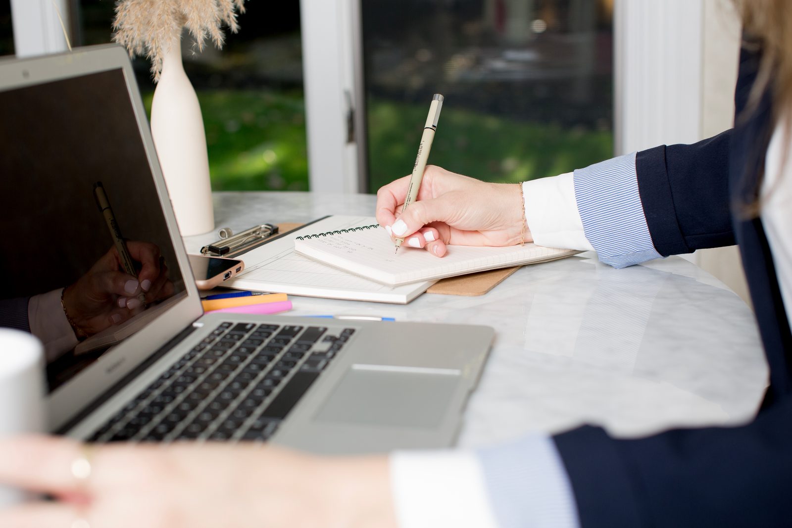Hands write in a notebook beside an open laptop on a marble table.