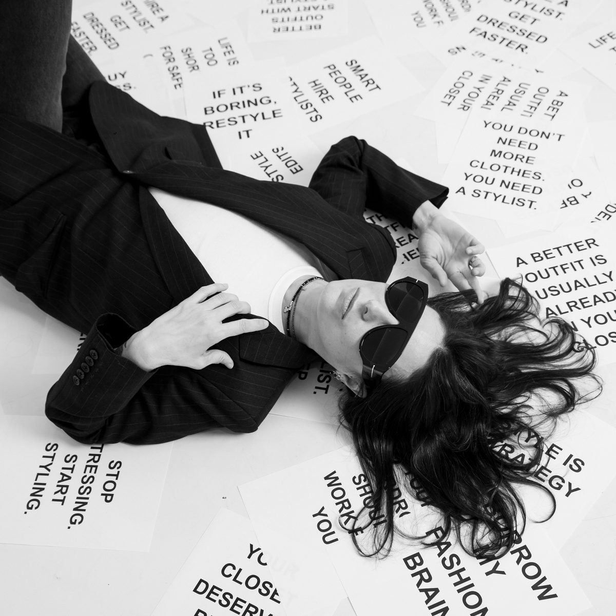 A person in a dark suit and dark sunglasses lies on a floor covered in printed text cards, in black and white.