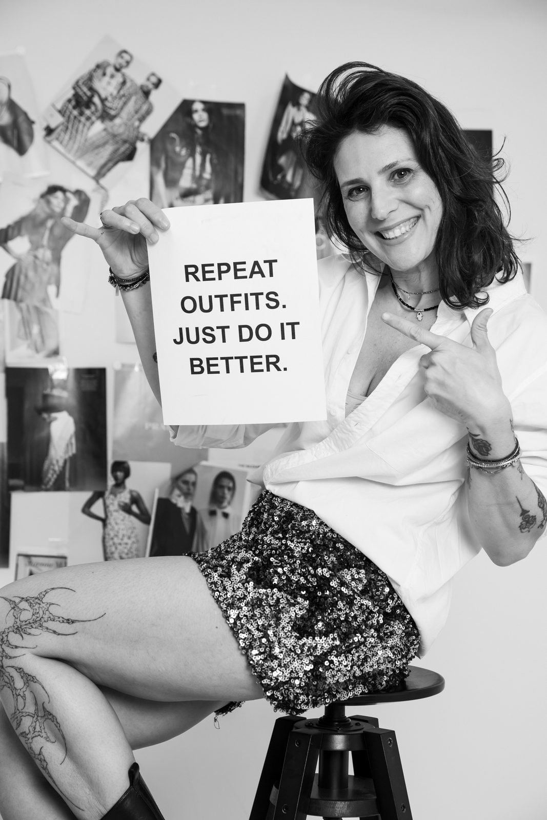 A smiling woman on a stool holds up a card reading Repeat outfits, just do it better, in front of a wall of photographs, in black and white.