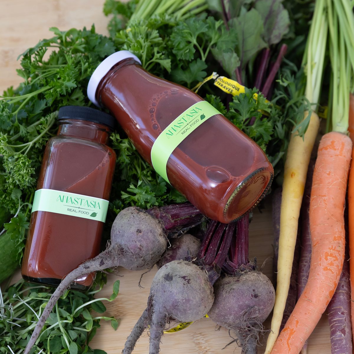 Two bottles of a deep red drink lie among beets, carrots and fresh herbs.
