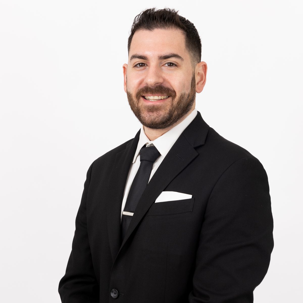 Corporate headshot of a smiling man with a dark beard in a black suit, white shirt and black tie against a white background.