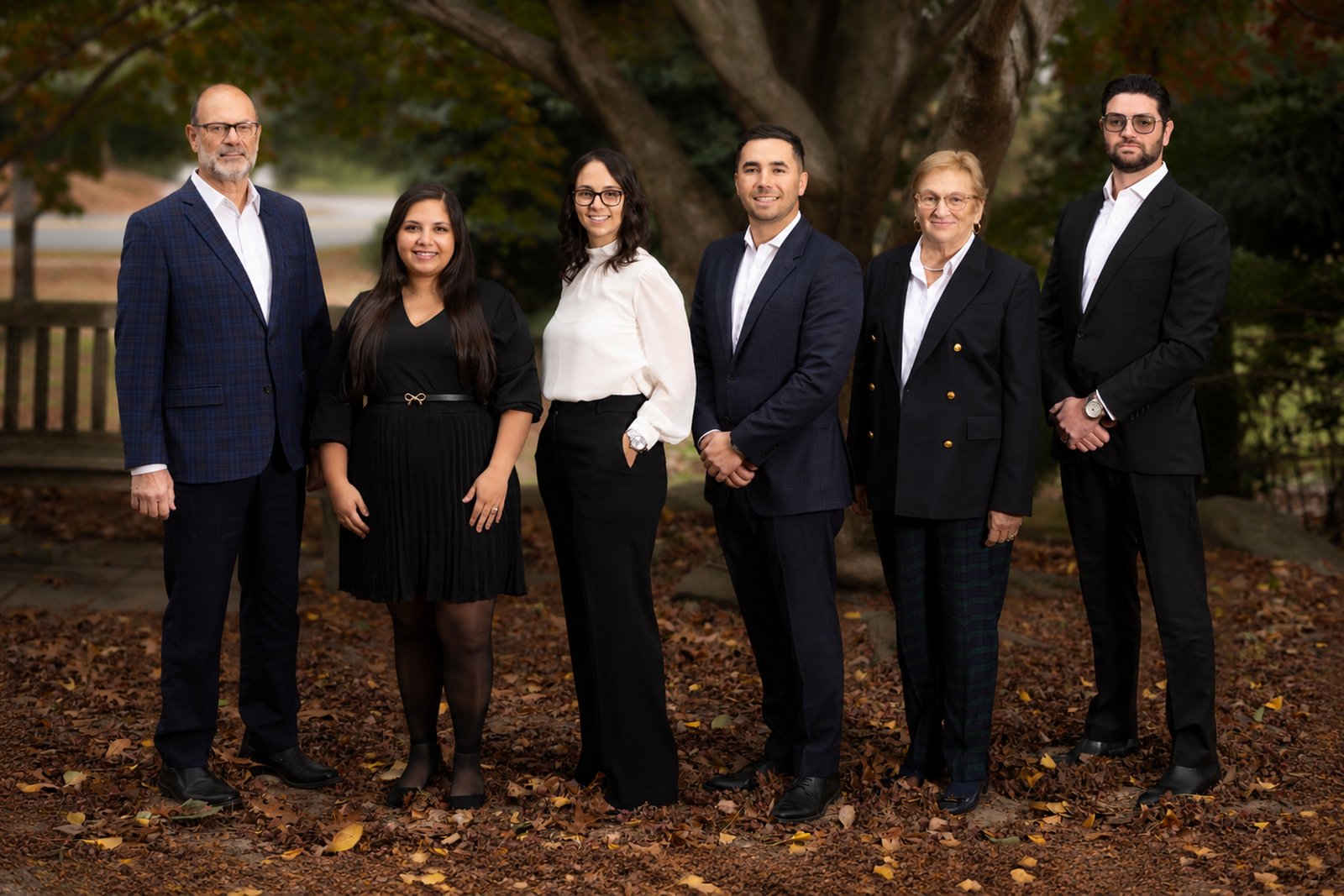 Six people in business attire stand together outdoors on autumn leaves in front of a large tree.