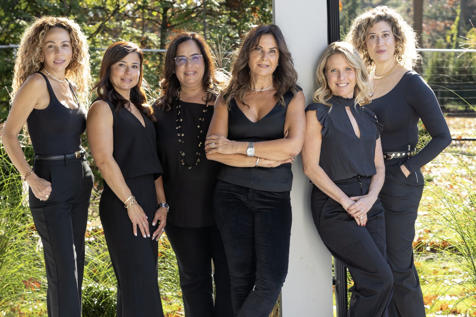 Six team members in black outfits lean against a white pillar outdoors for a relaxed group portrait.
