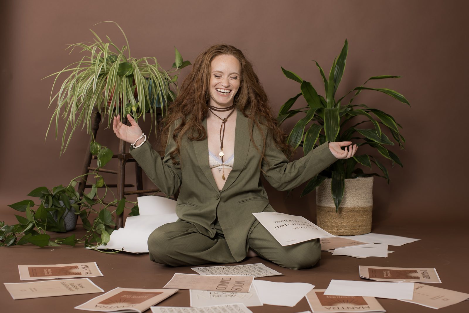 A woman with long red hair in an olive suit laughs, seated among plants and printed cards on a brown studio backdrop.