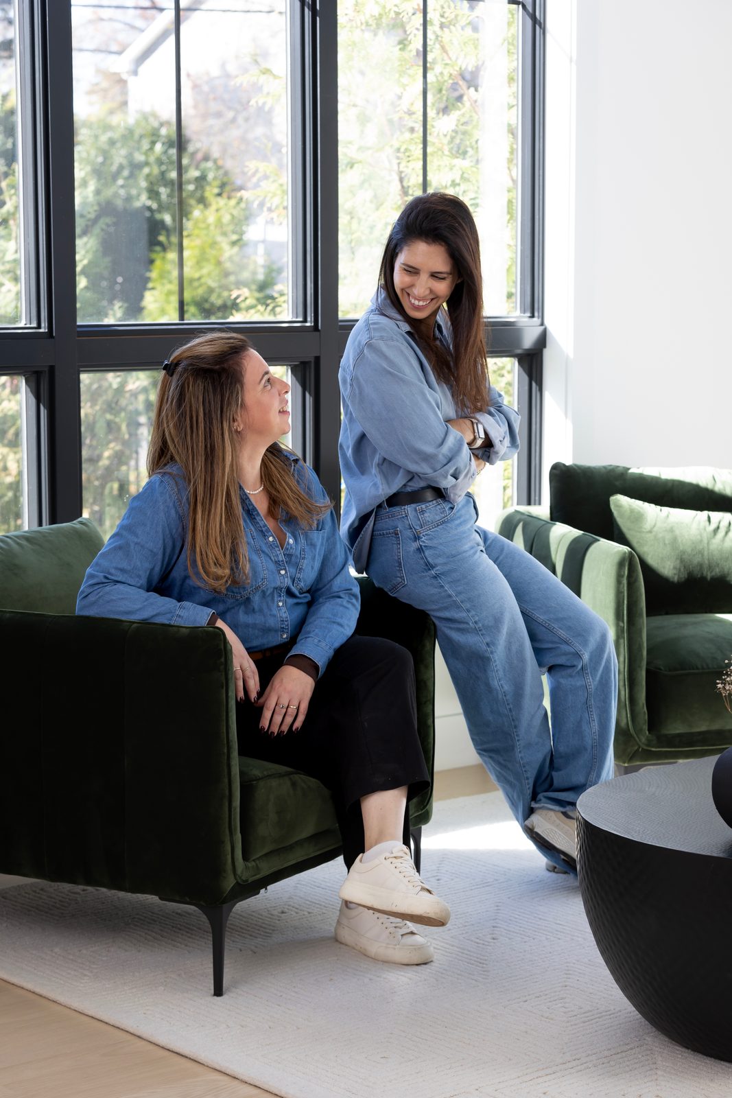 Two women in denim talk and laugh by tall windows, one seated in a green armchair and one perched on its arm.