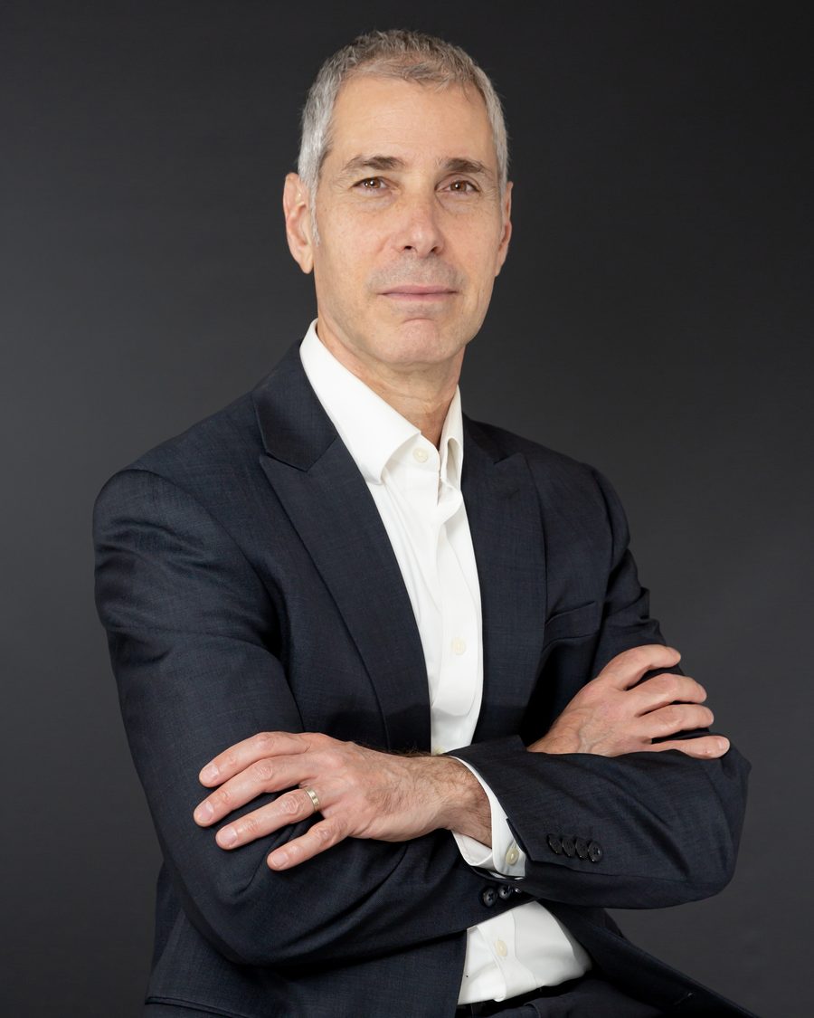 A silver-haired man in a dark suit with arms crossed against a charcoal backdrop.