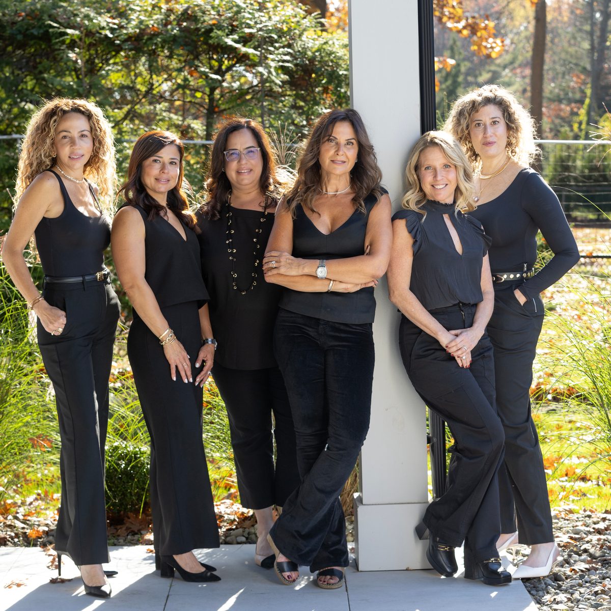 Six women in black outfits stand together by a white column outdoors, with autumn trees behind them.