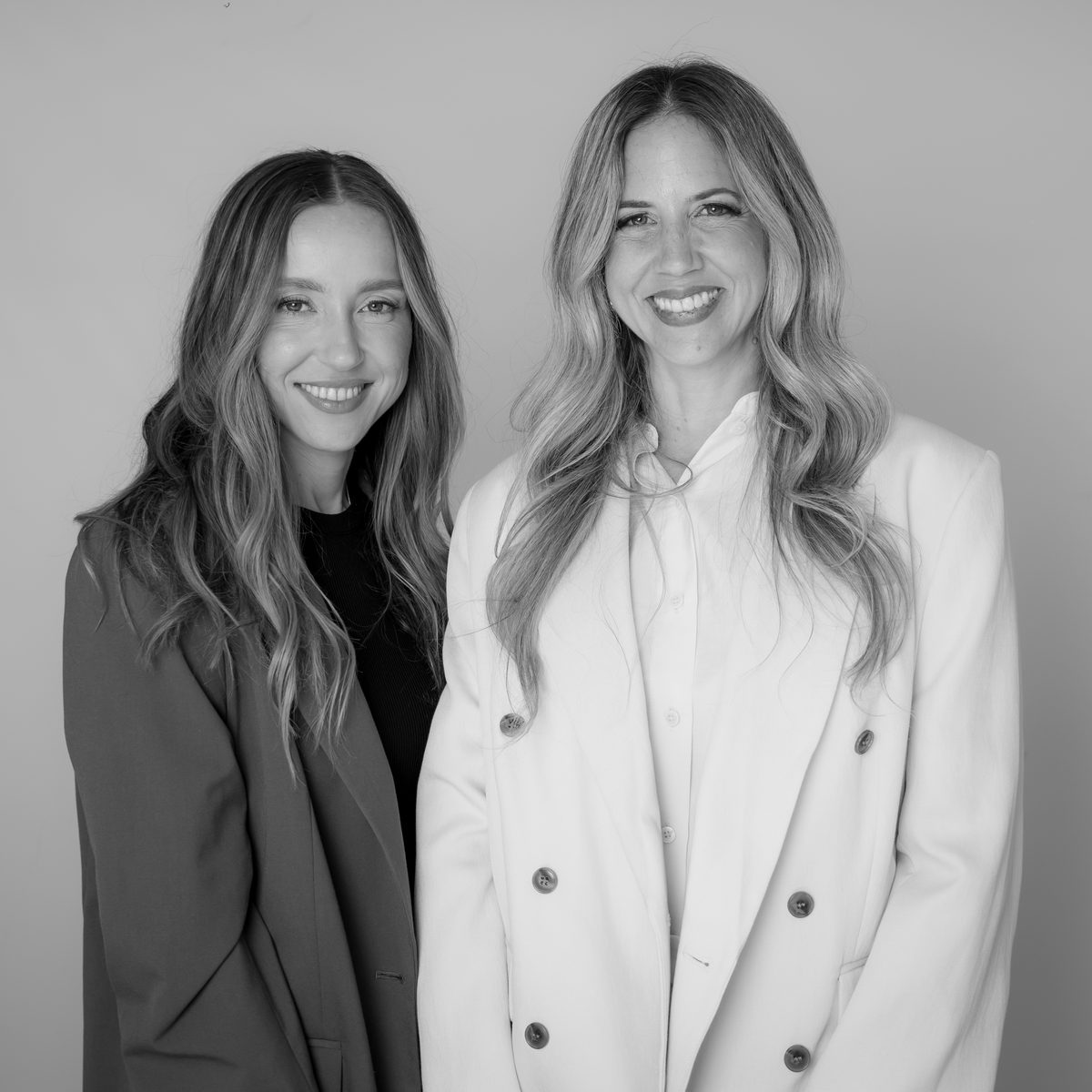 Two smiling women with long hair stand side by side, one in a dark coat and one in a white blazer, in black and white.