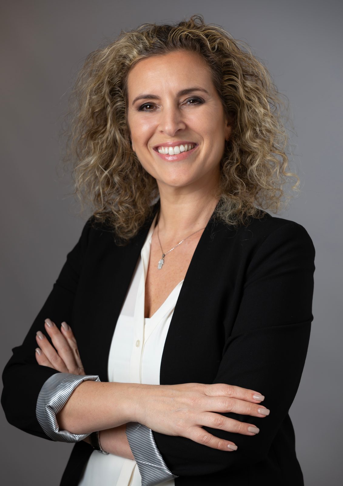 A smiling woman with curly hair in a black blazer and white blouse, arms crossed, against a gray studio backdrop.