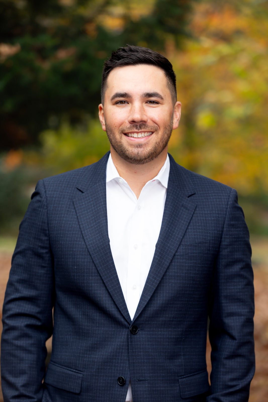 A smiling man in a navy checked blazer and white shirt, photographed outdoors against autumn trees.