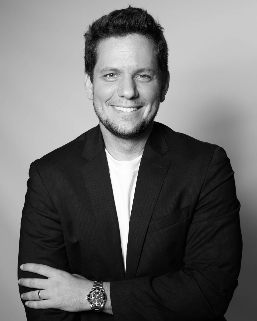 A smiling man with short dark hair in a dark blazer over a white T-shirt, arms folded, in black and white.
