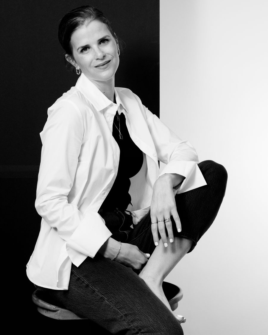 A smiling woman in a white shirt and dark jeans sits on a stool against a dark and light backdrop, in black and white.
