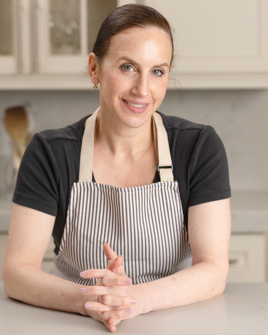 A smiling woman in a striped apron leans on a kitchen counter with her hands clasped.